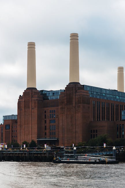 The image shows the historic Battersea Power Station, a large red-brick building with multiple tall smokestacks rising into a cloudy sky. In the foreground, a waterway is visible with a docked barge a