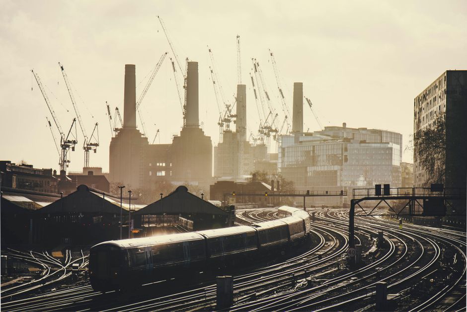 The image shows the historic Battersea Power Station, a large red-brick building with multiple tall smokestacks rising into a cloudy sky. In the foreground, a waterway is visible with a docked barge a