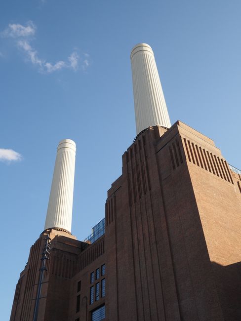 A low-angle view of a large industrial building with red brick walls and multiple rectangular windows on the left side. A modern glass balcony runs along the building's edge, supported by a concrete s